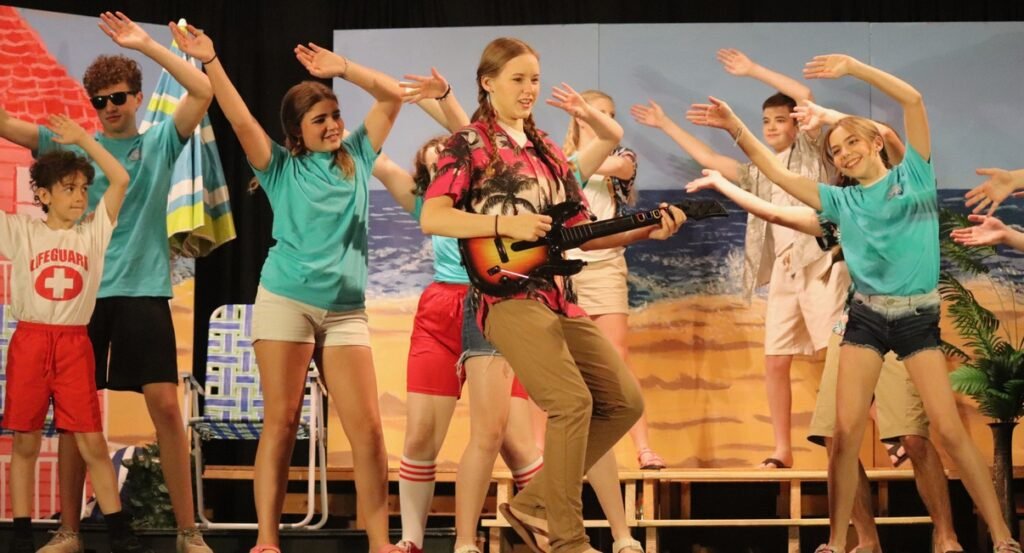 Cast members in beach-themed costumes on stage in Much Ado About Nothing